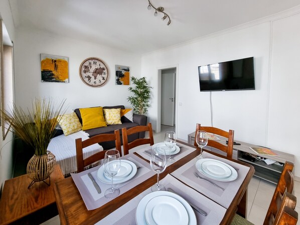 A dining area with a wooden table set for four, a sofa with yellow cushions, a clock, and a TV mounted on the wall.