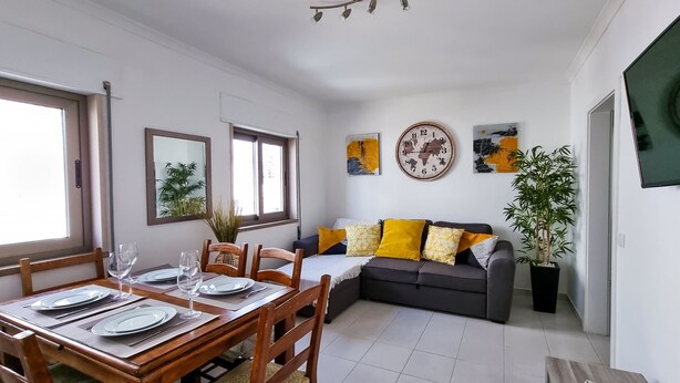 Un coin repas avec une table en bois dressée pour quatre personnes, un canapé avec des coussins jaunes et une plante en pot.