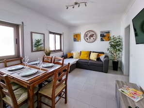 A dining area with a wooden table set for four, a sofa with yellow cushions, and a potted plant.