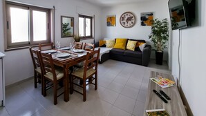 Un coin repas avec une table en bois dressée pour quatre personnes, un canapé avec des coussins jaunes et un téléviseur fixé au mur.