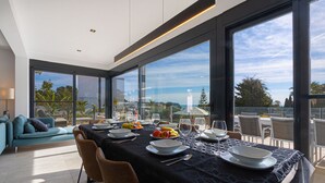 Uma sala de jantar moderna com uma mesa grande posta para refeição, grandes janelas com vista para o exterior e uma área de estar confortável com um sofá.