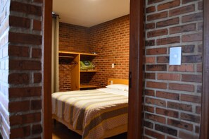 A small, cozy room with a brick wall, a wooden bed, and a shelf with a bag and a green object.