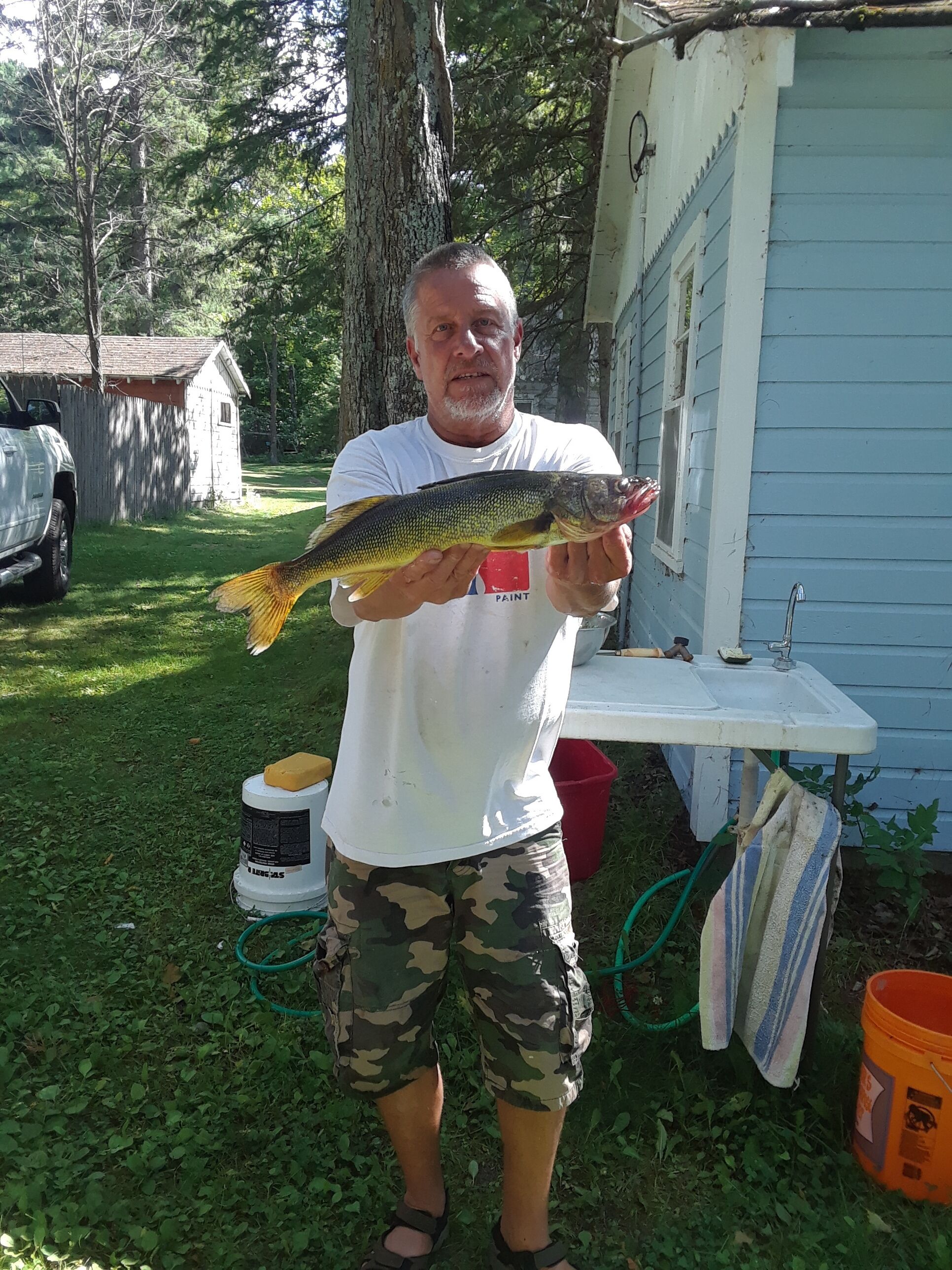 A person holding a fish outdoors near a house with a sink and a red bucket.