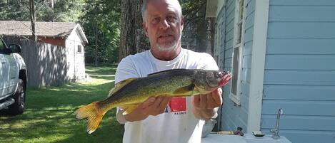 A person holding a fish outdoors near a house with a sink and a red bucket.
