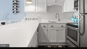 Eine Küche mit weißen Schränken, einem Edelstahlkühlschrank und einem eingebauten Backofen. An der Wand ist ein Weinregal angebracht und ein Schild mit der Aufschrift 'BEACH HOUSE'.