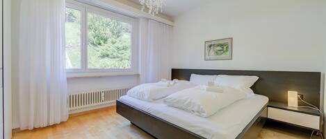 Un dormitorio con una cama grande, una mesita de noche, una araña, una ventana con cortinas y un radiador.