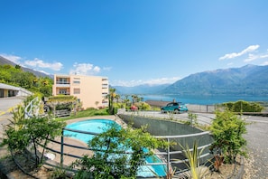 En swimmingpool omgiven av grönska, en byggnad med flera balkonger och utsikt över berg och en sjö.