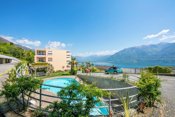 Ein Schwimmbad, umgeben von Grünflächen, ein Gebäude mit mehreren Balkonen und Blick auf Berge und einen See.