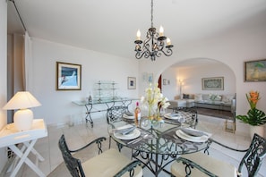 A dining area with a glass table, metal chairs, a chandelier, and a white sofa in the background.