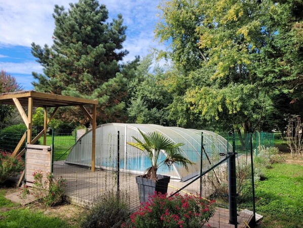 A greenhouse structure with a wooden pergola nearby, surrounded by a fenced garden area with various plants and trees.