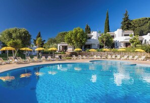 Una piscina con sillones de descanso y sombrillas amarillas, rodeada de edificios blancos y vegetación.