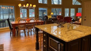 A spacious kitchen with a large island, wooden dining table, and chairs, large windows with a view of trees, and a ceiling fan. - Reduced Spring rates! On Lake Oconee with Sunset Views, 2 Masters (Eatonton)