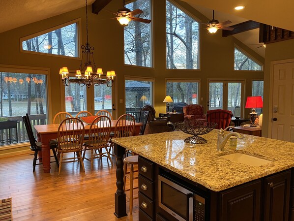 A spacious kitchen with a large island, wooden dining table, and chairs, large windows with a view of trees, and a ceiling fan.