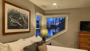 A bedroom with a large bed, a nightstand with a lamp, a dresser, a framed picture on the wall, and a window with a view of trees and a sunset. - Reduced Spring rates! On Lake Oconee with Sunset Views, 2 Masters (Eatonton)