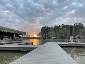 A wooden dock extending into a calm body of water, with a covered seating area and a view of a sunset over a forested area.