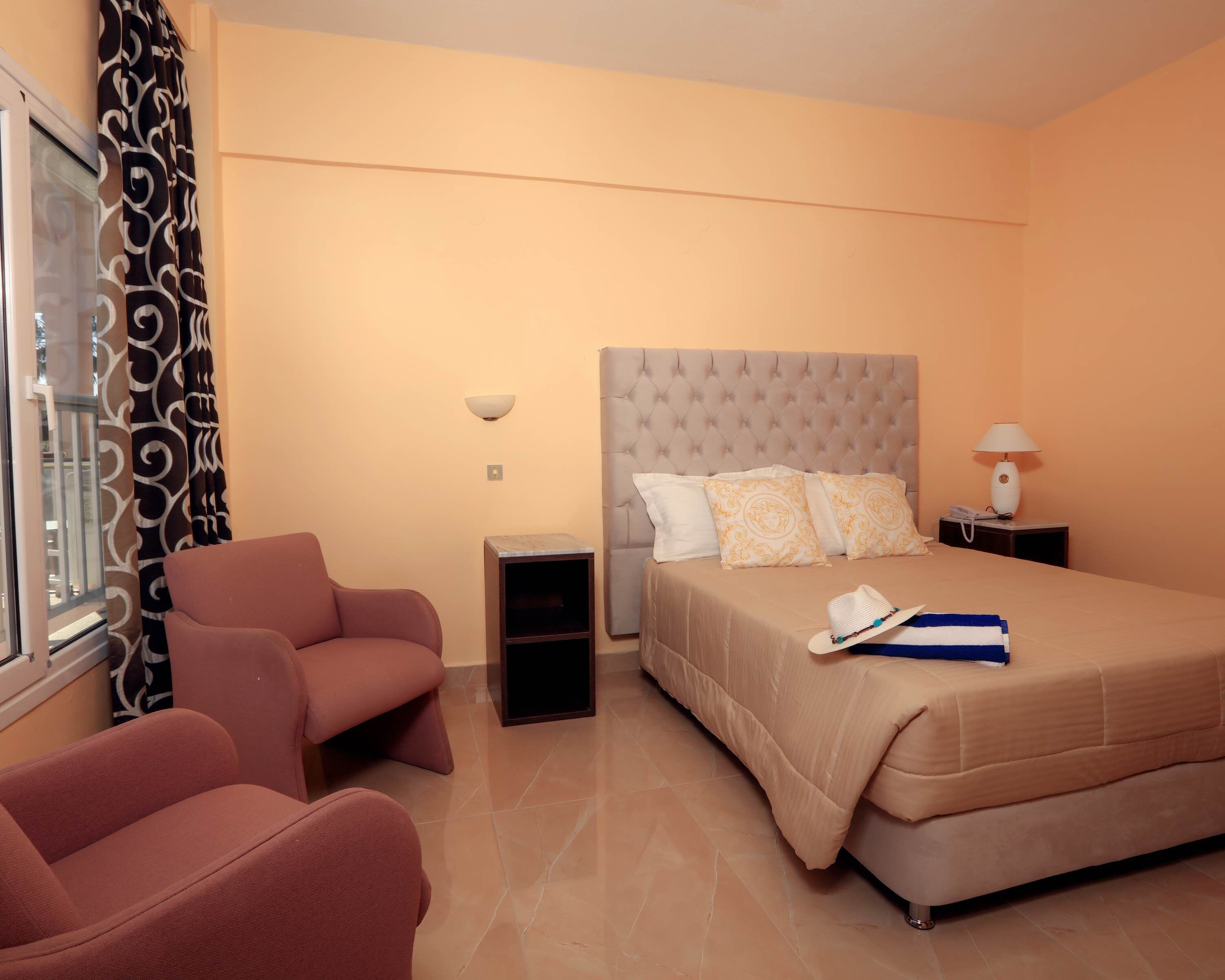Une chambre d’hôtel avec un lit, une chaise, une table de chevet, une lampe et une fenêtre avec des rideaux.