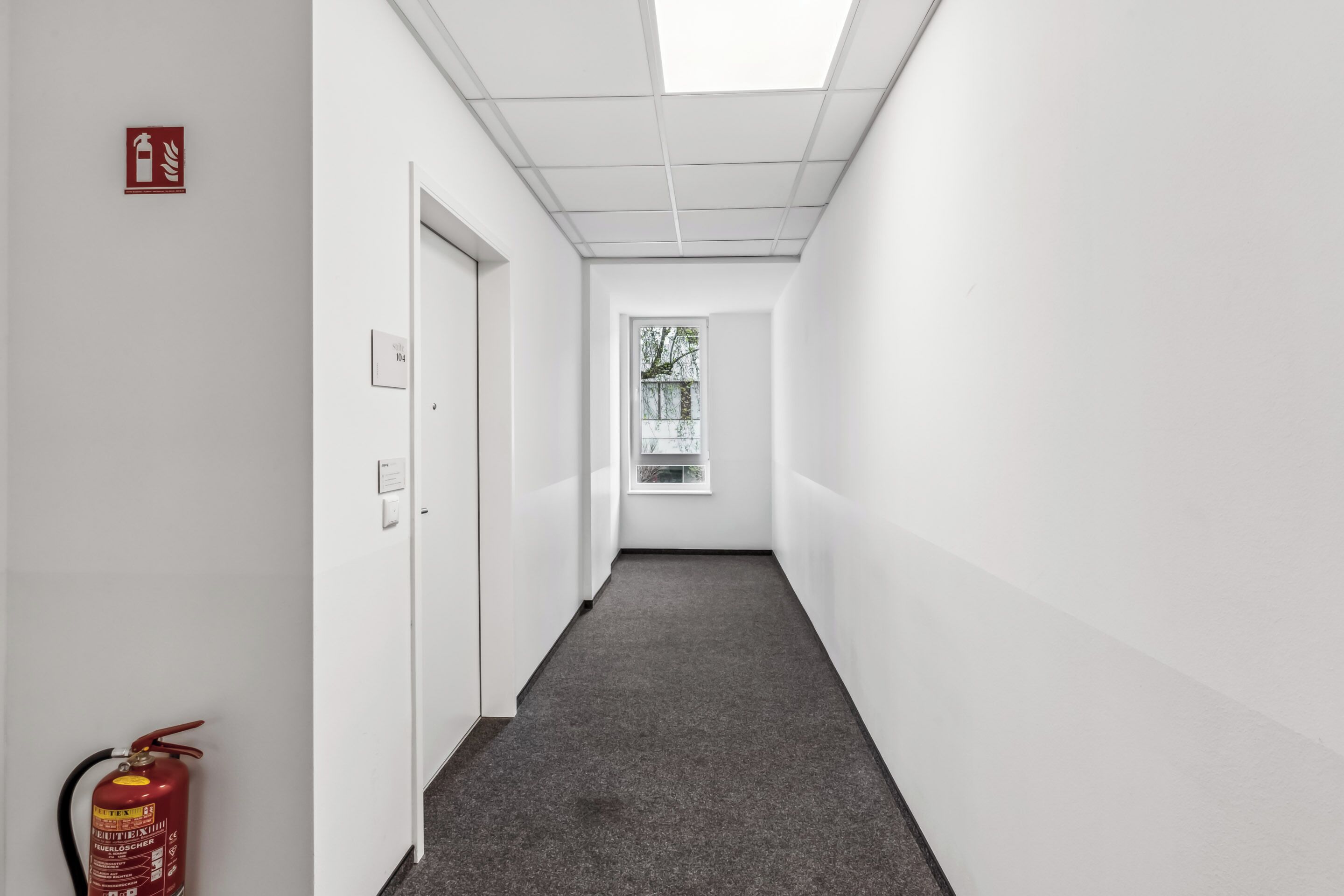 Un couloir étroit et bien éclairé, avec un extincteur sur le côté gauche, une porte au fond et une fenêtre à l’extrémité.