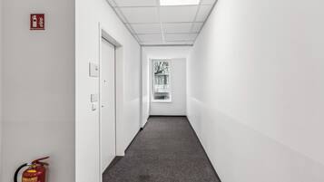 Un couloir étroit et bien éclairé, avec un extincteur sur le côté gauche, une porte au fond et une fenêtre à l’extrémité.