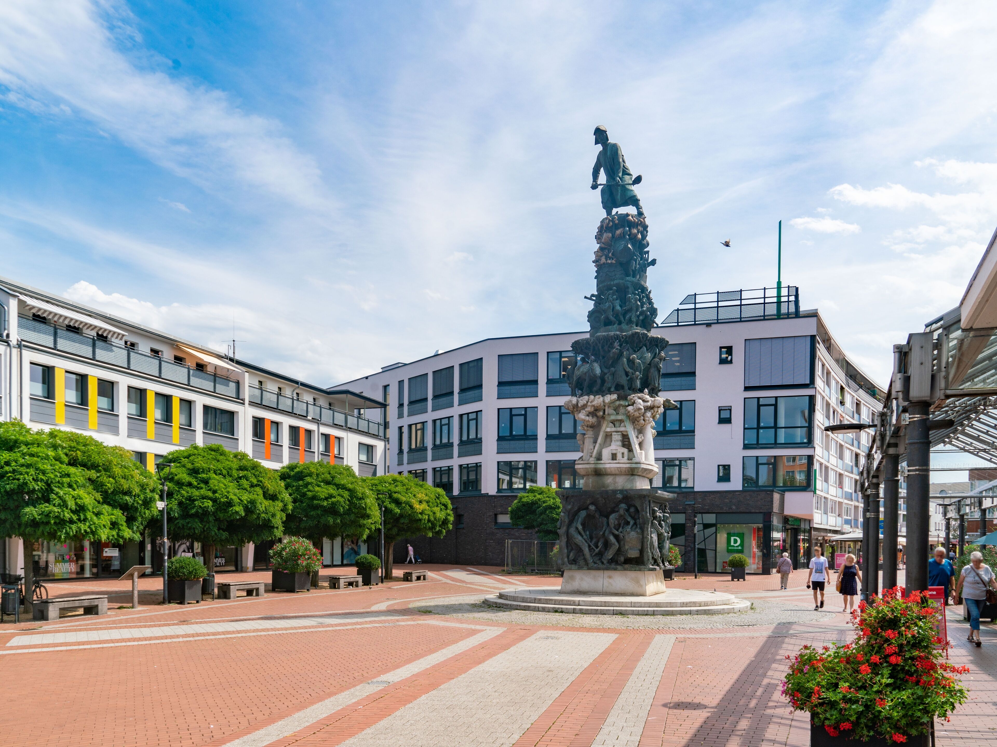 A public square with a central statue, surrounded by modern buildings and greenery.