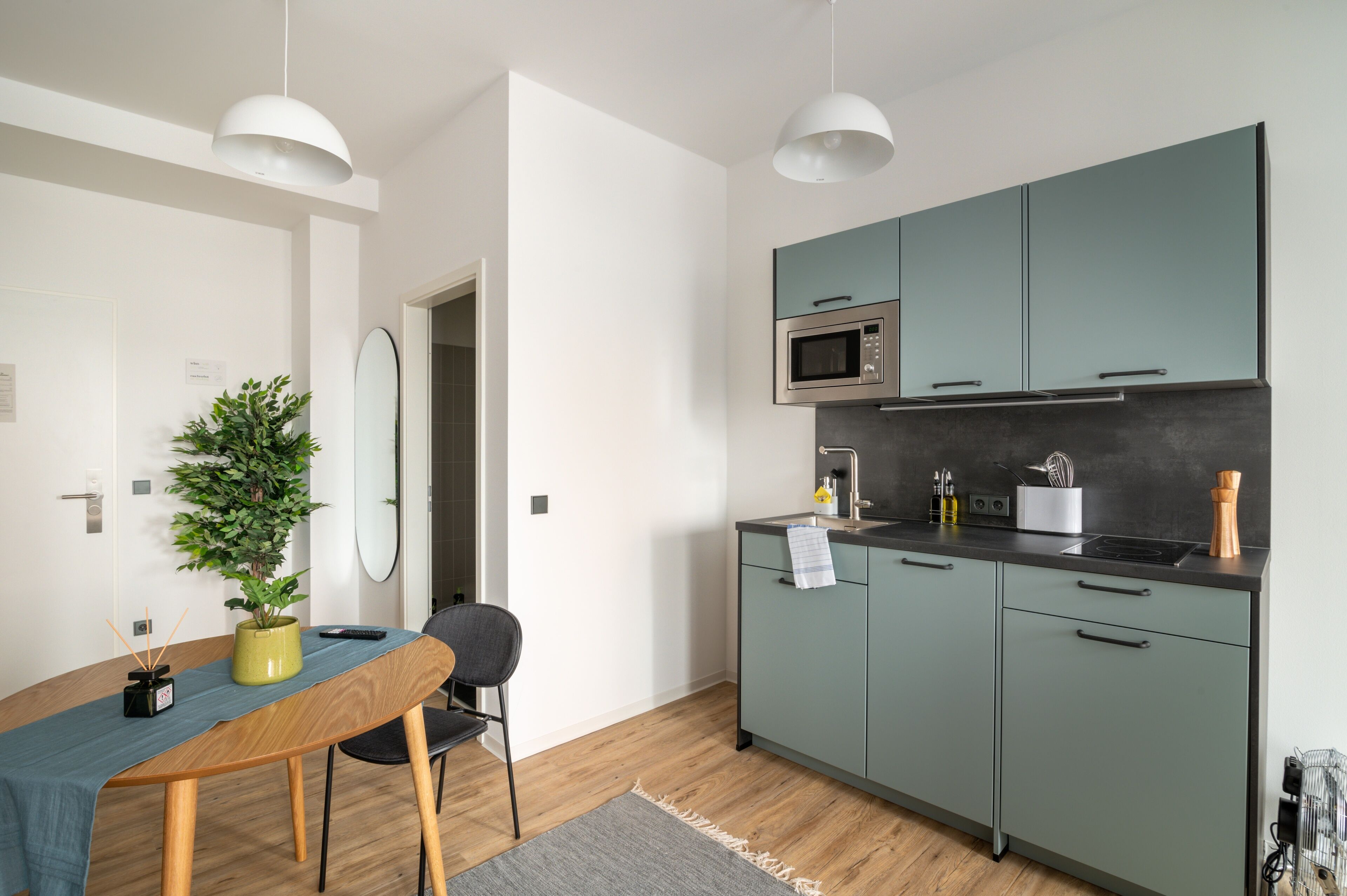 Een moderne keuken met blauwgroene kastjes, een magnetron en een kookplaat. Er staat een eettafel met stoelen en een potplant.