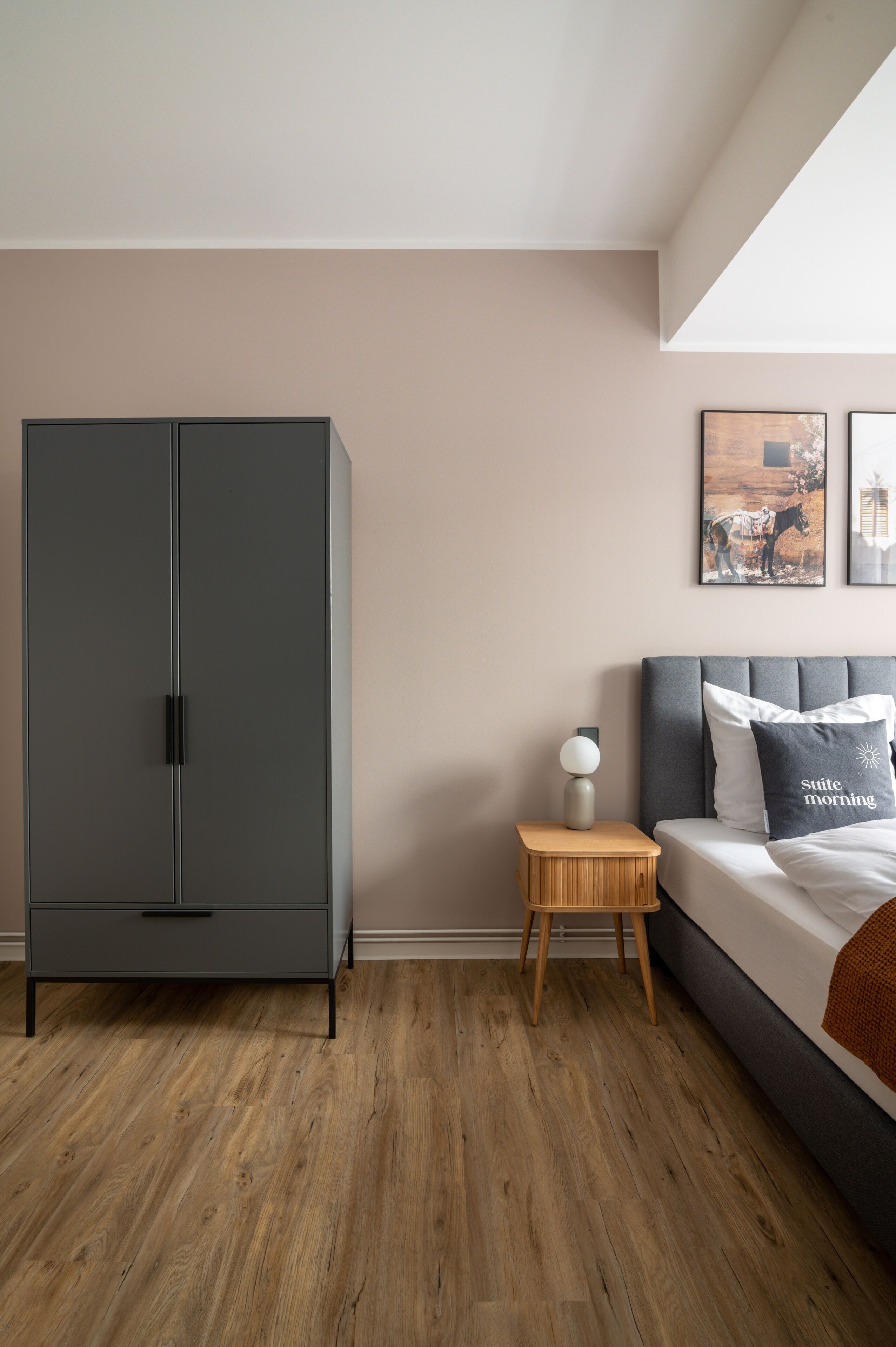 A bedroom with a grey wardrobe, a bedside table with a lamp, a bed with a grey headboard, and two framed pictures on the wall.