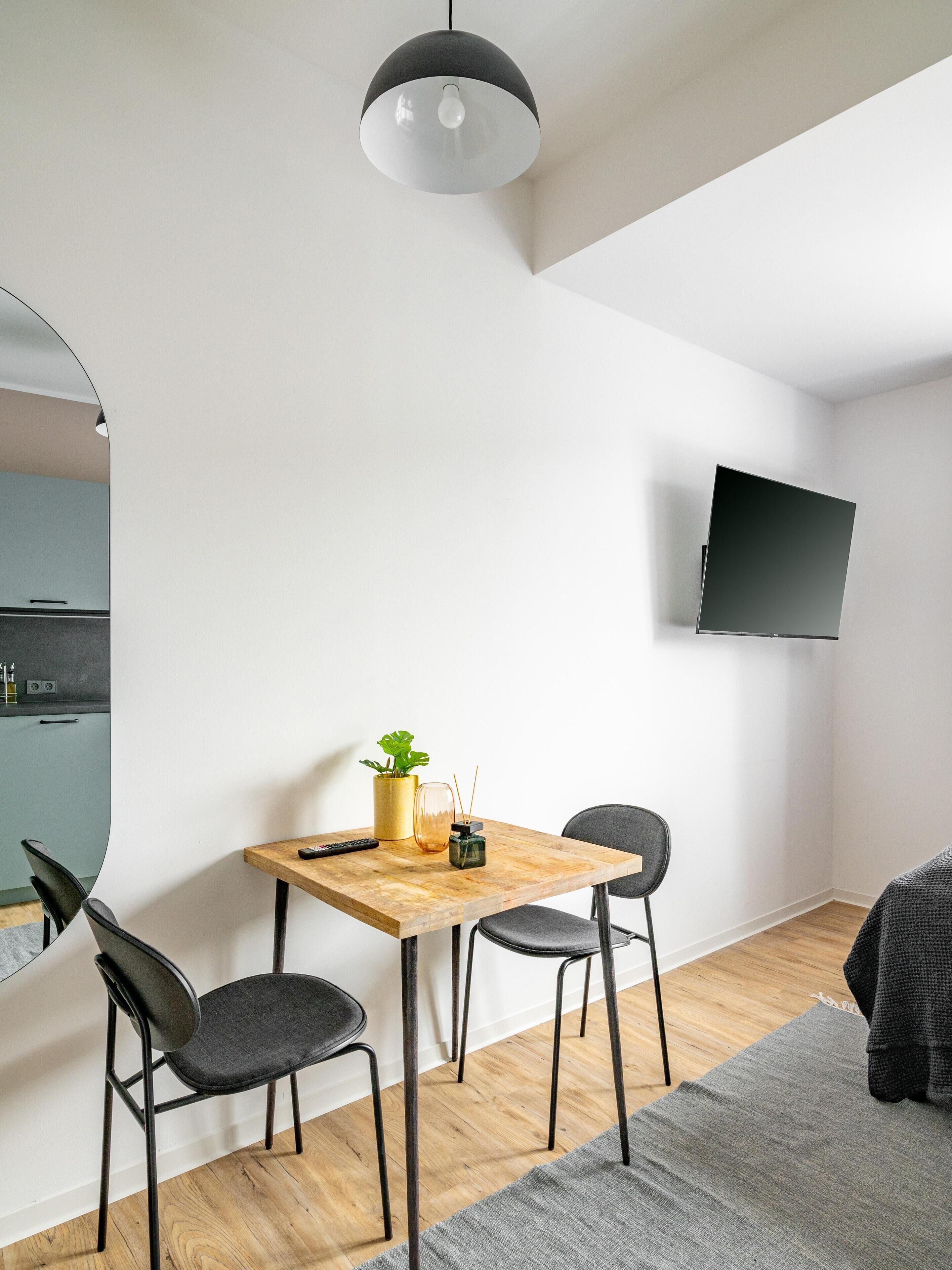 A modern, minimalist living space with a wooden dining table, two chairs, a small plant, and a flat-screen TV mounted on the wall.