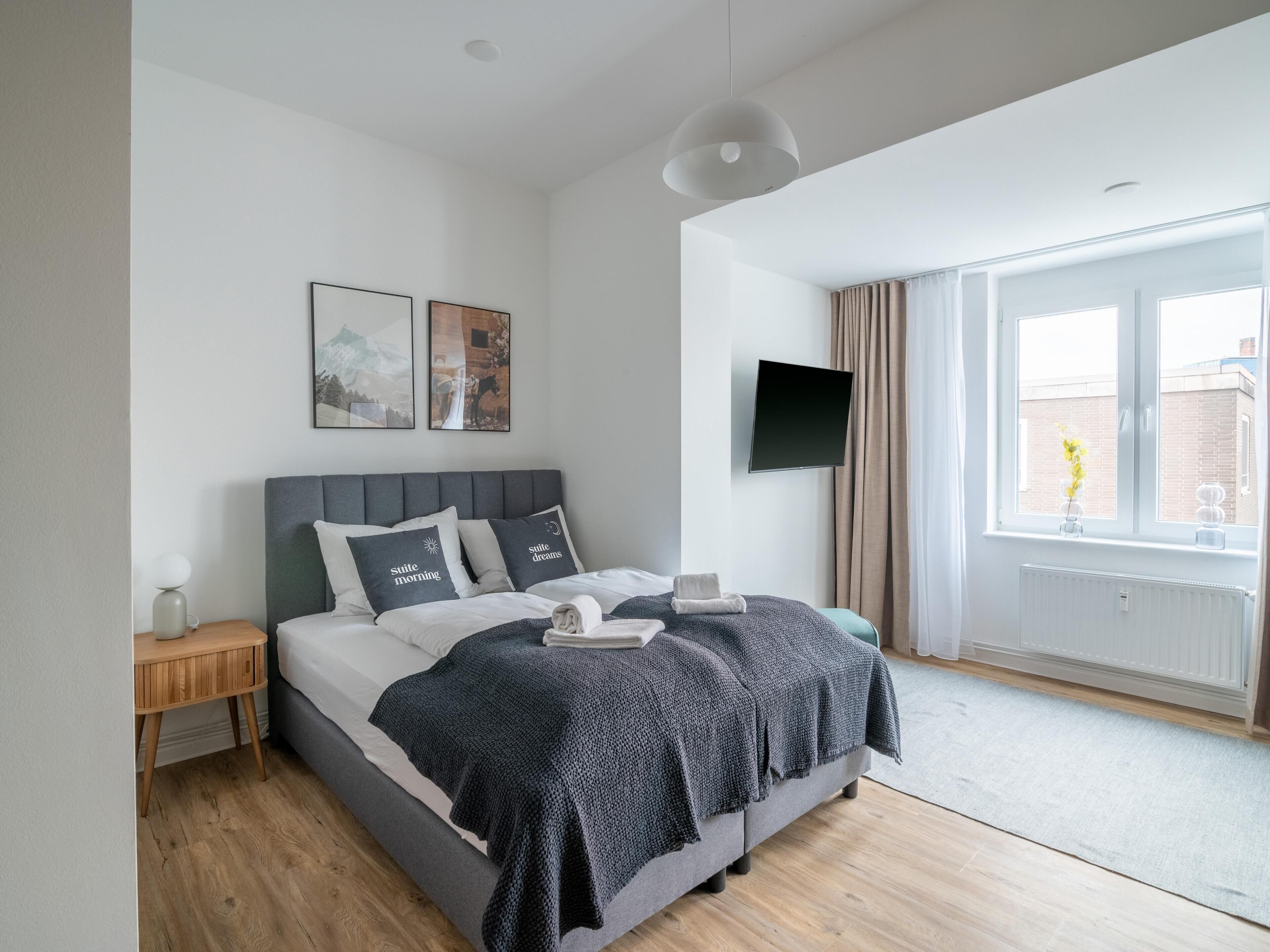 A modern bedroom with a large bed, a bedside table, a flat-screen TV, and a window with curtains.