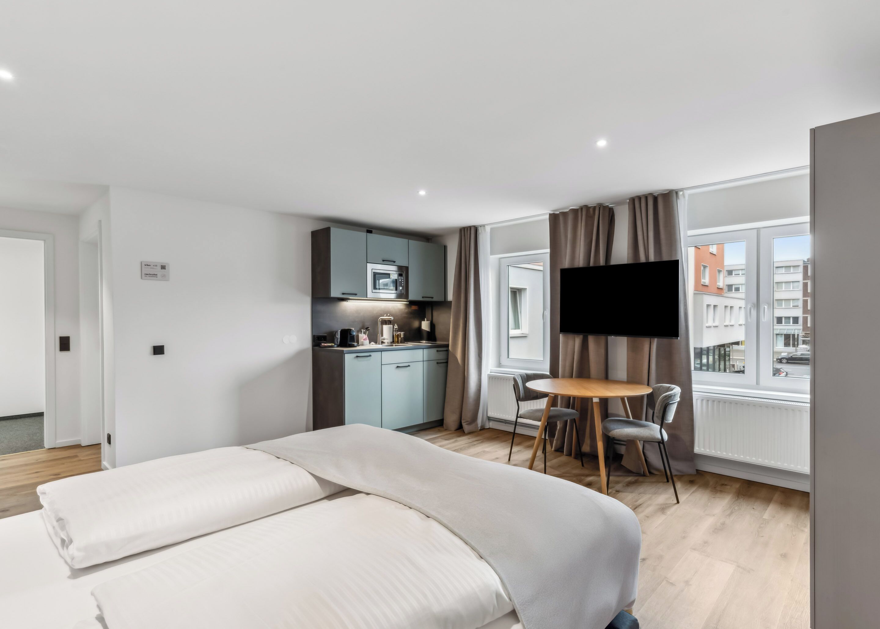 A modern hotel room with a bed, a dining table with chairs, a kitchenette, and a TV mounted on the wall.