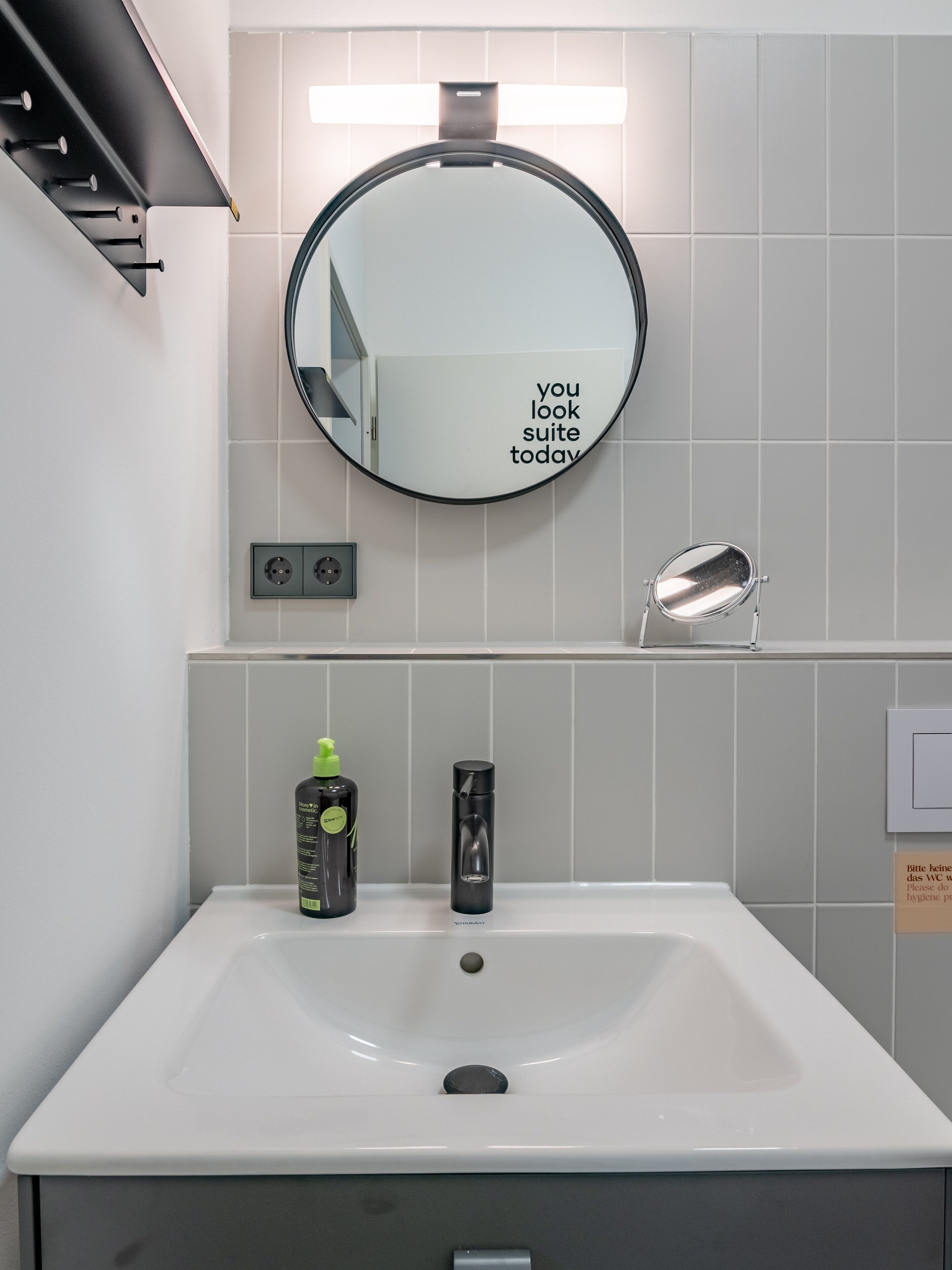 A modern bathroom with a white sink, a round mirror with a motivational quote, a handheld mirror, and toiletries on the counter.
