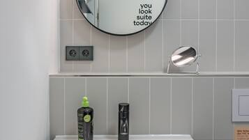 A modern bathroom with a white sink, a round mirror with a motivational quote, a handheld mirror, and toiletries on the counter.