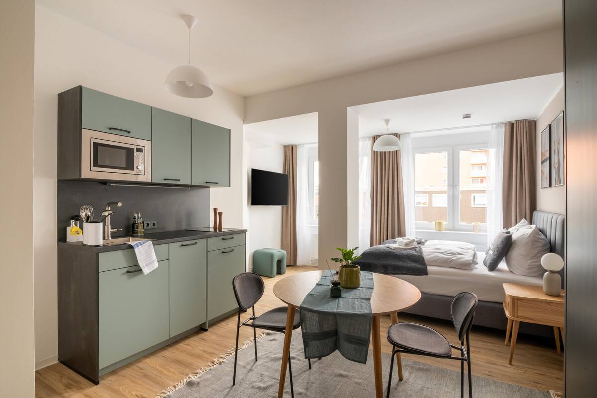 A compact hotel room with a kitchenette, a dining area, a bed with a view, and a TV.