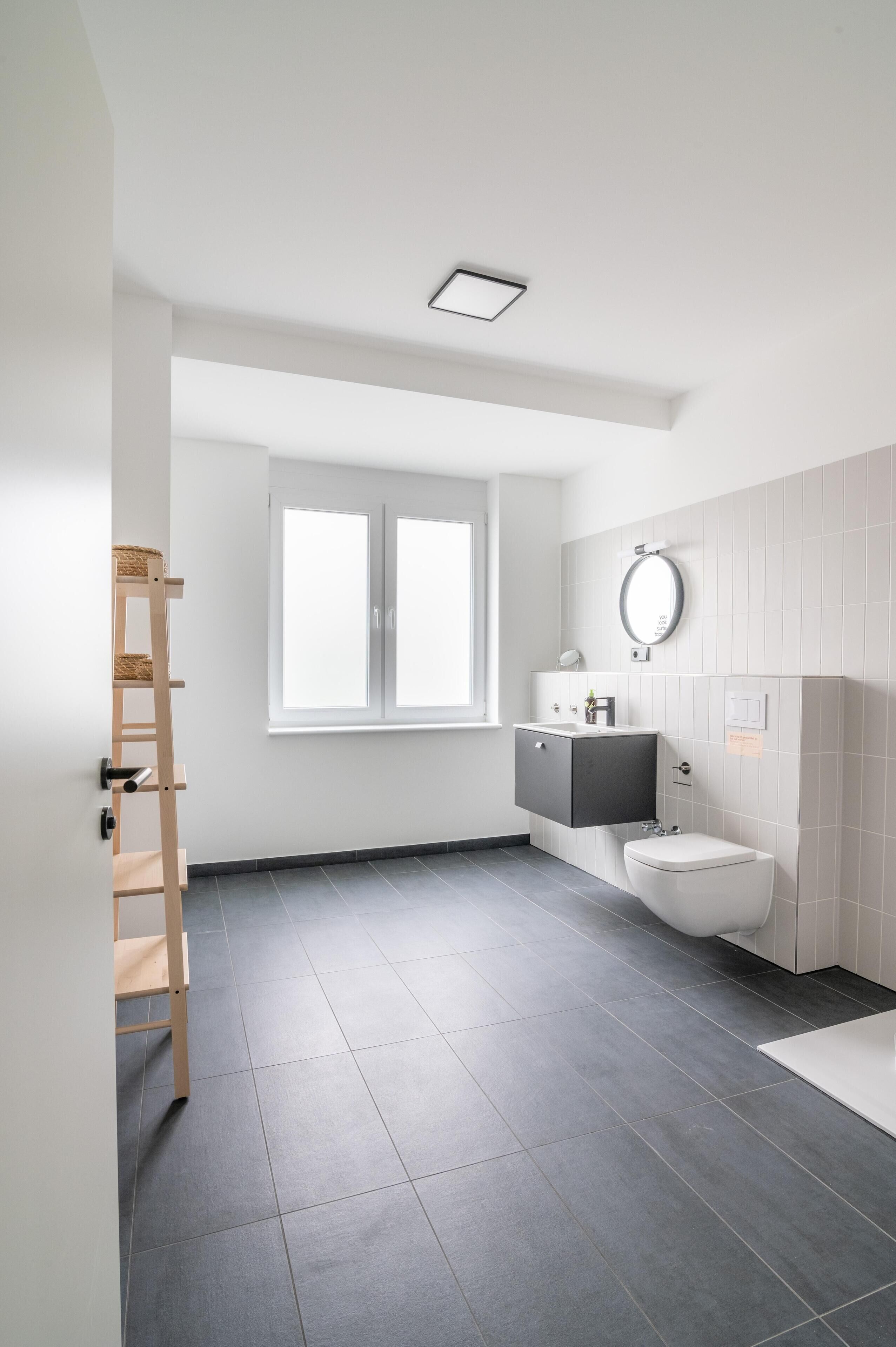 Ein modernes Badezimmer mit grau gefliestem Boden, einer weißen Toilette, einem dunkelgrauen Waschtisch und einem Fenster, das für natürliches Licht sorgt.