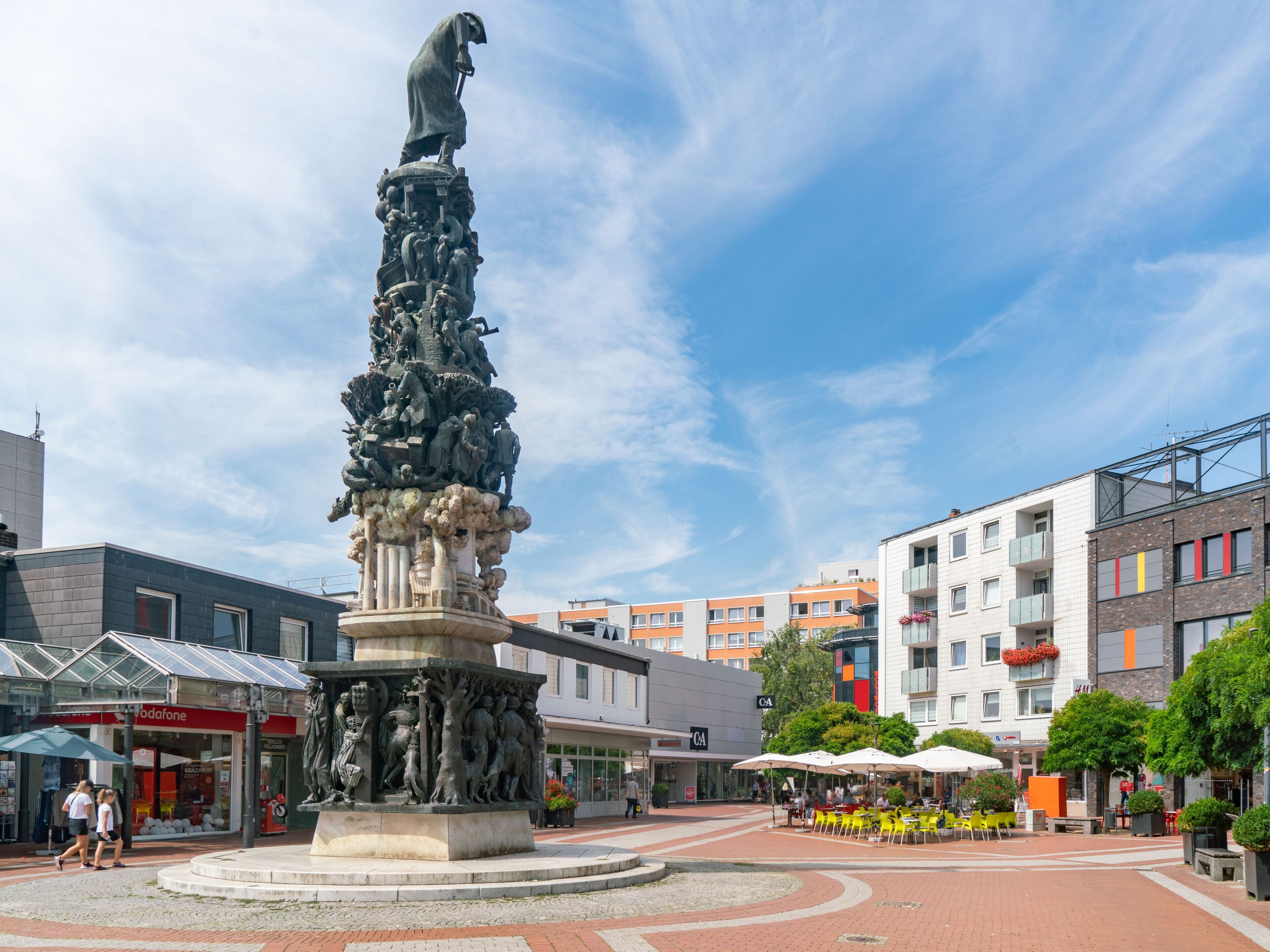 Una plaza pública con un monumento central, edificios circundantes y zonas de estar al aire libre.