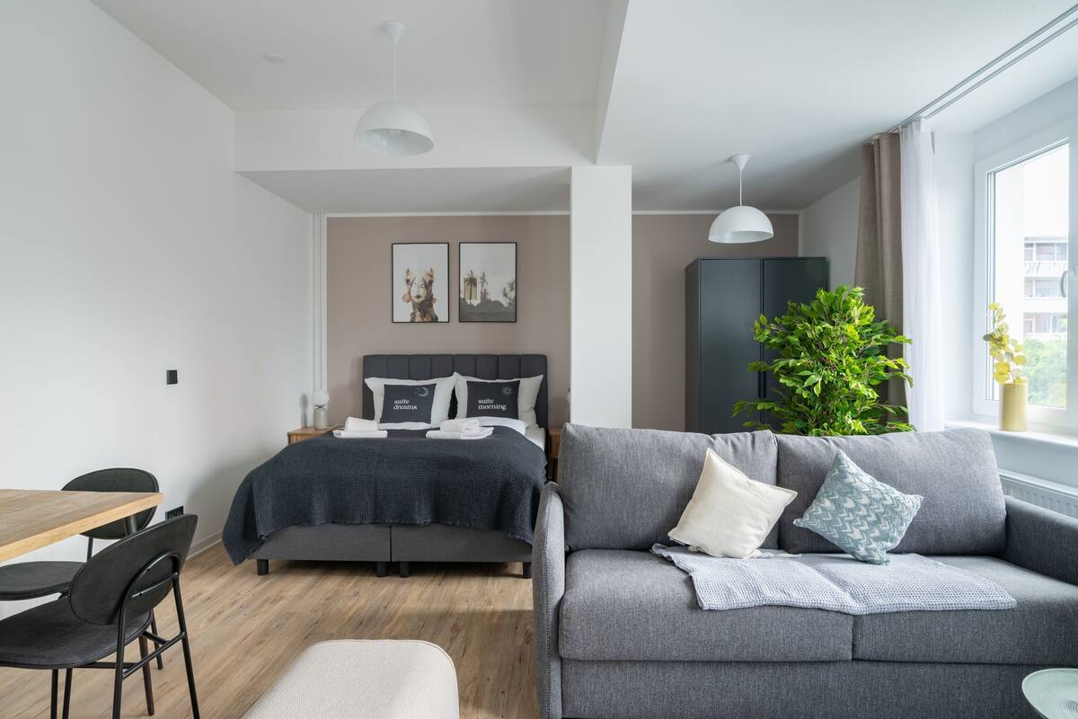 A modern bedroom with a bed, a grey sofa, a dining table, chairs, and a large window.