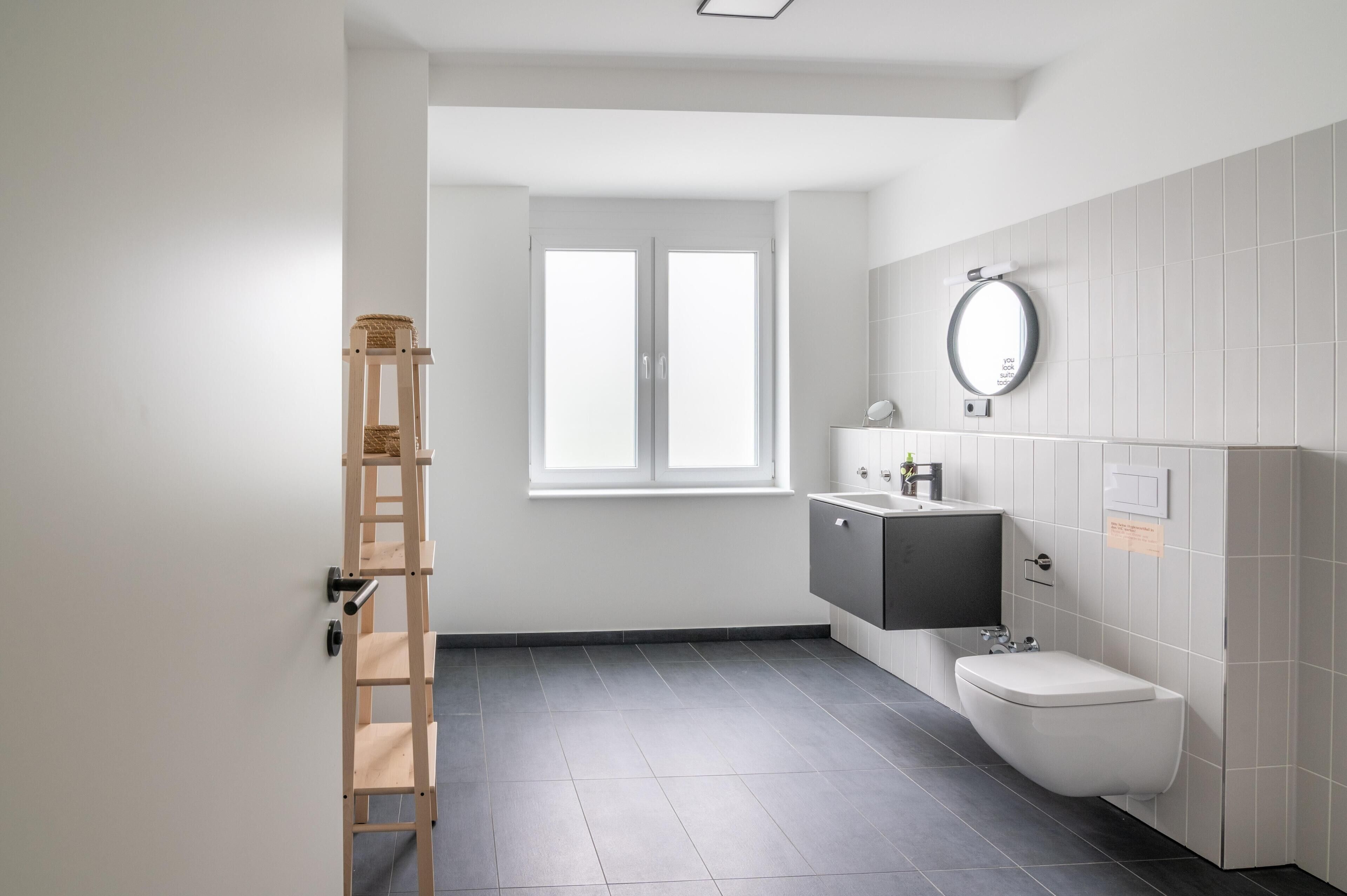 A modern bathroom with a toilet, sink, and mirror. There is a window providing natural light and a ladder against the wall.