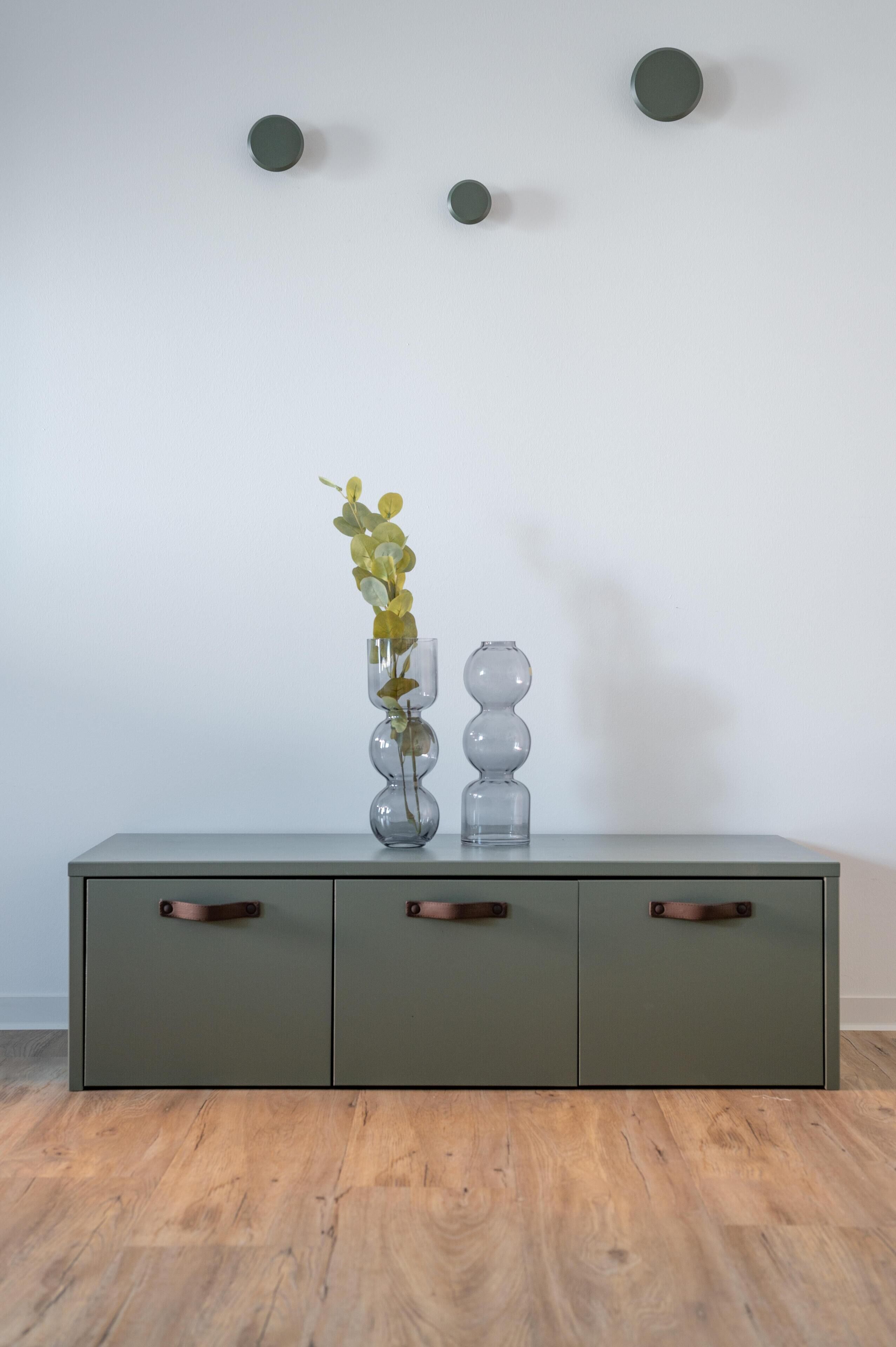 Ein grauer Schrank mit drei Schubladen und einer Vase mit grünen Blättern darauf.