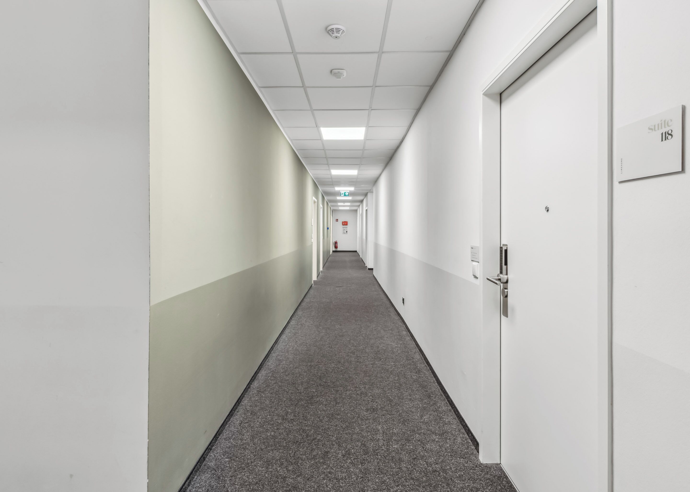 A long, narrow hallway with white walls and a grey carpet. There are doors on both sides, and a sign indicating 'suite 118' on the right wall.