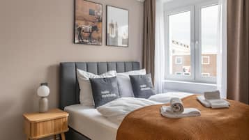 Une chambre à coucher moderne avec un grand lit, une table de chevet, une fenêtre avec des rideaux et des œuvres d’art accrochées au mur.