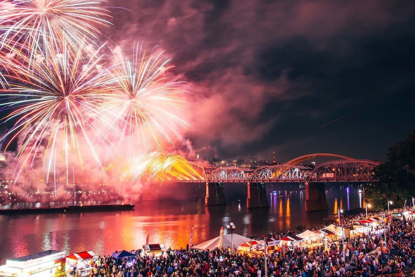 A nighttime outdoor event with fireworks, a crowd, and a bridge.