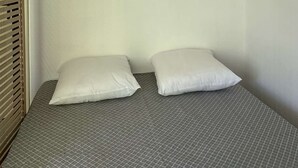 Quarto com cama, duas almofadas, prateleira pequena com uma planta, luminária na parede e um espelho decorativo.