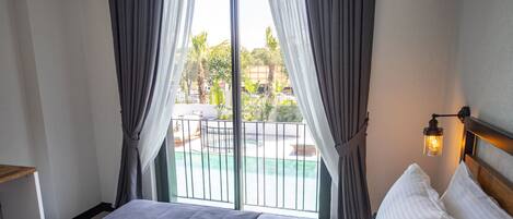 A hotel room with a bed, a nightstand, a window with curtains, and a view of a swimming pool and greenery outside.