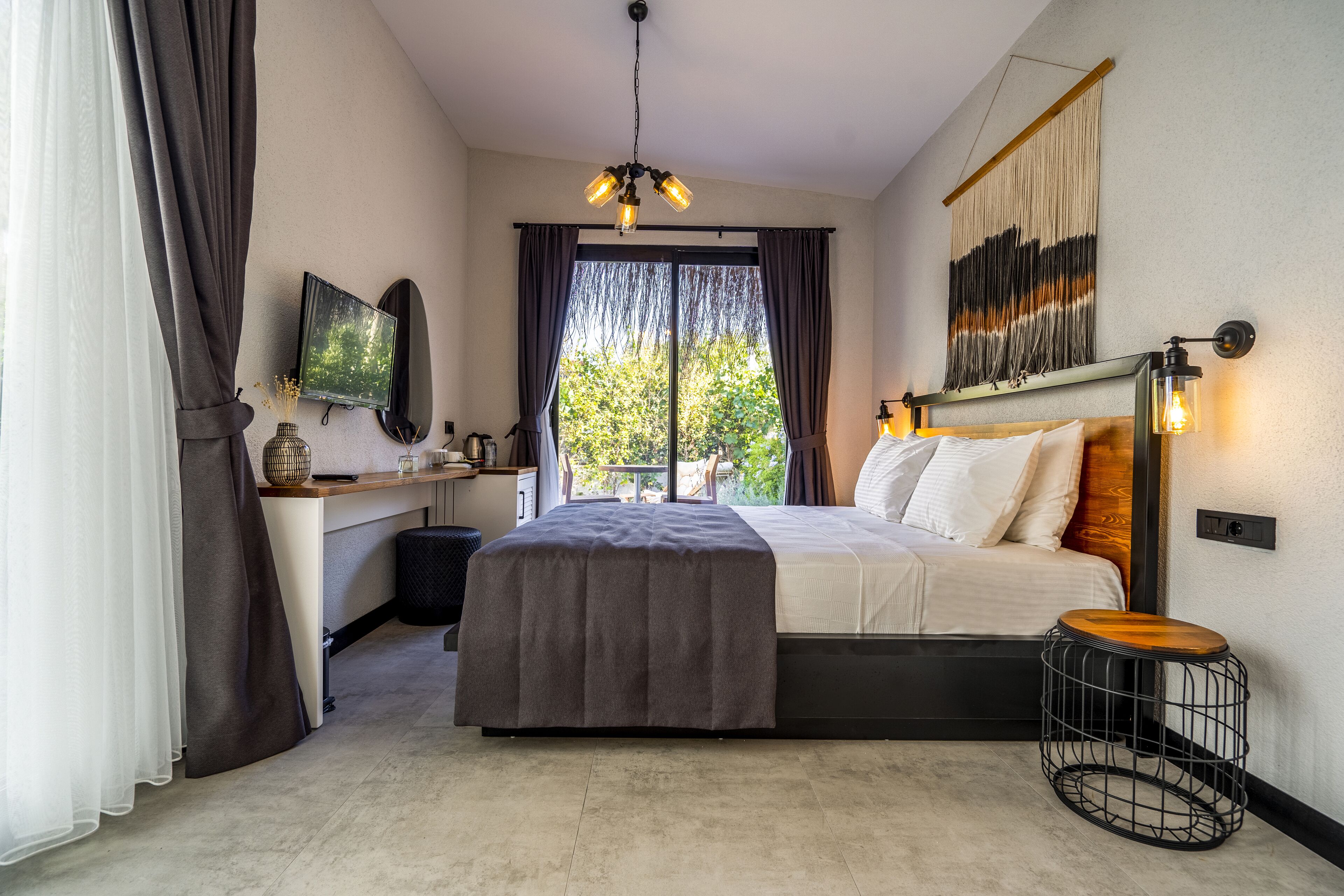 A modern bedroom with a large bed, a bedside table, a small stool, a TV, and a view of greenery outside.