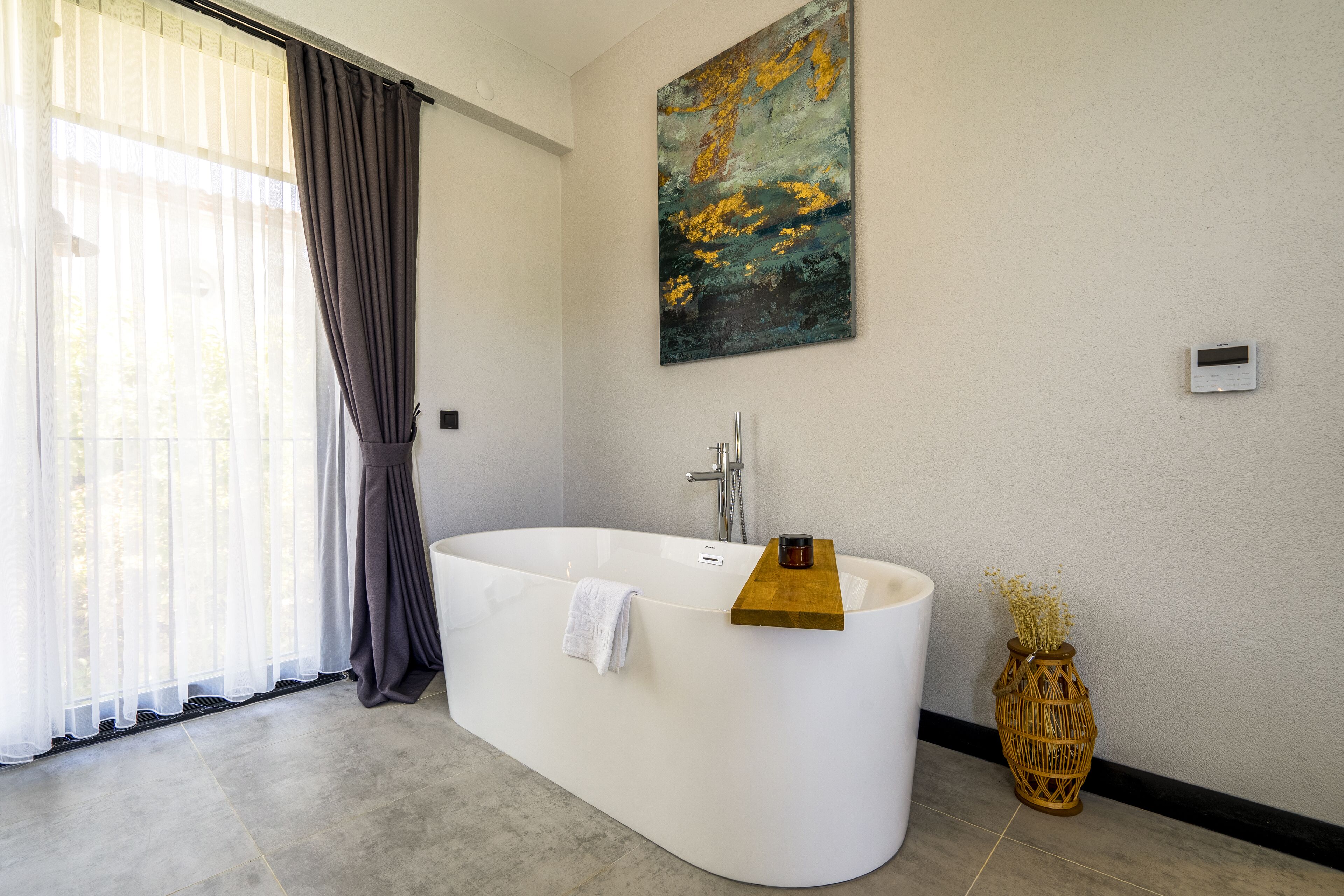 Een moderne badkamer met een vrijstaand bad, een groot raam met transparante gordijnen en een kunstwerk aan de muur.