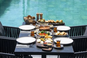 Mesa de comedor dispuesta con diversos platos y bebidas, con vista a una piscina.