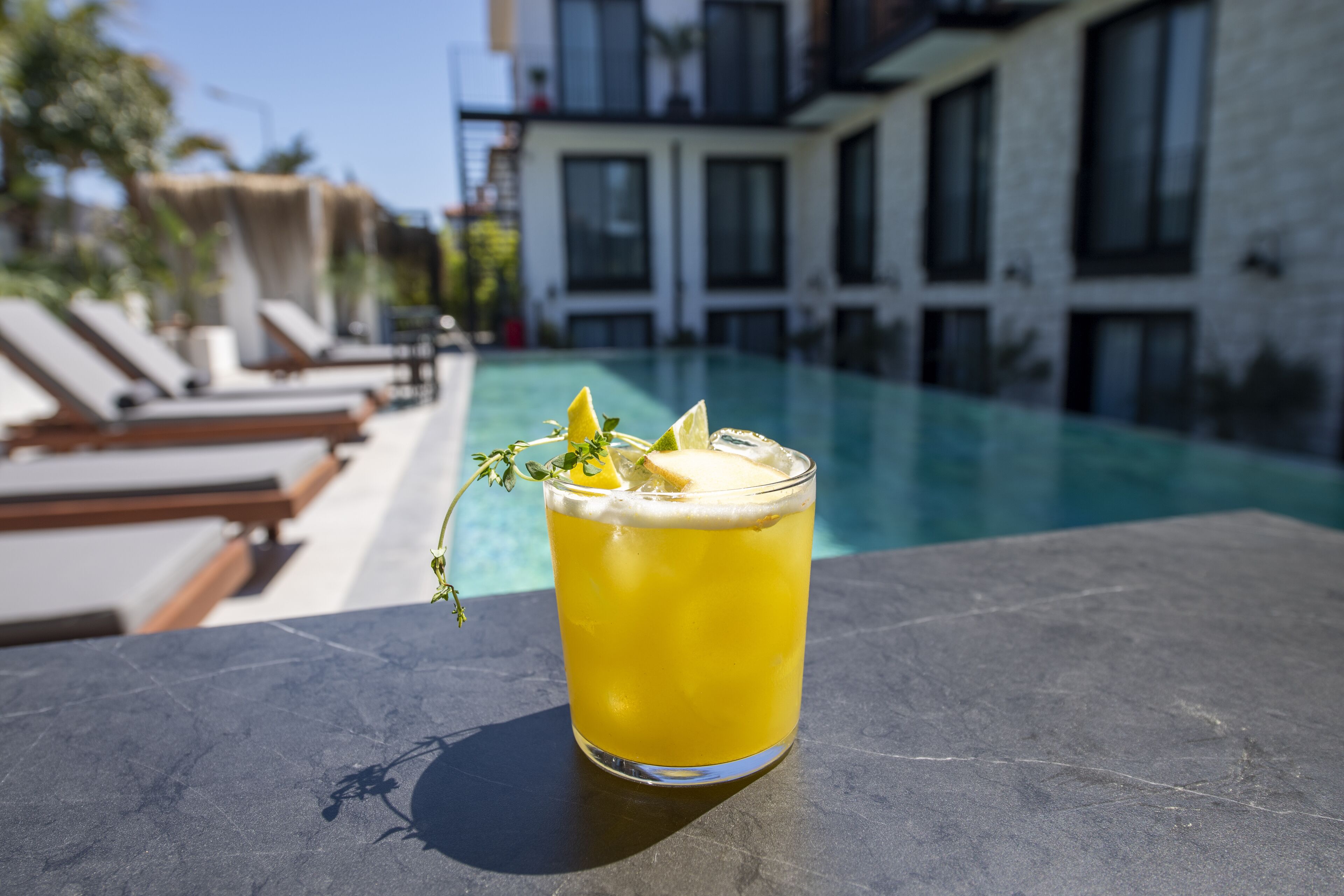 Un verre d’une boisson jaune, garni d’une tranche de citron et de quelques feuilles de menthe, posé sur une surface en pierre, au bord d’une piscine.