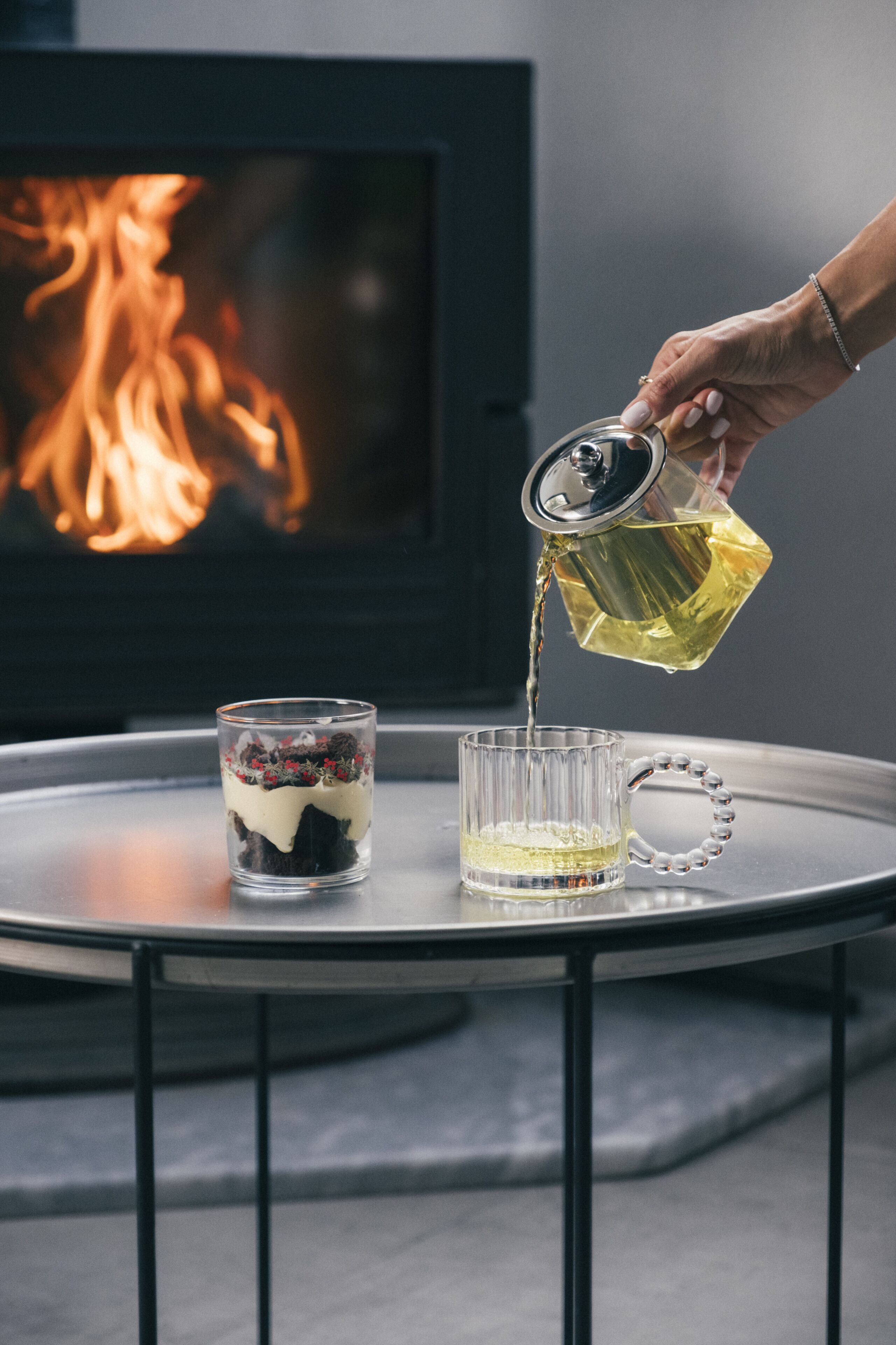 A hand pouring tea into a glass on a round table with a fireplace in the background.