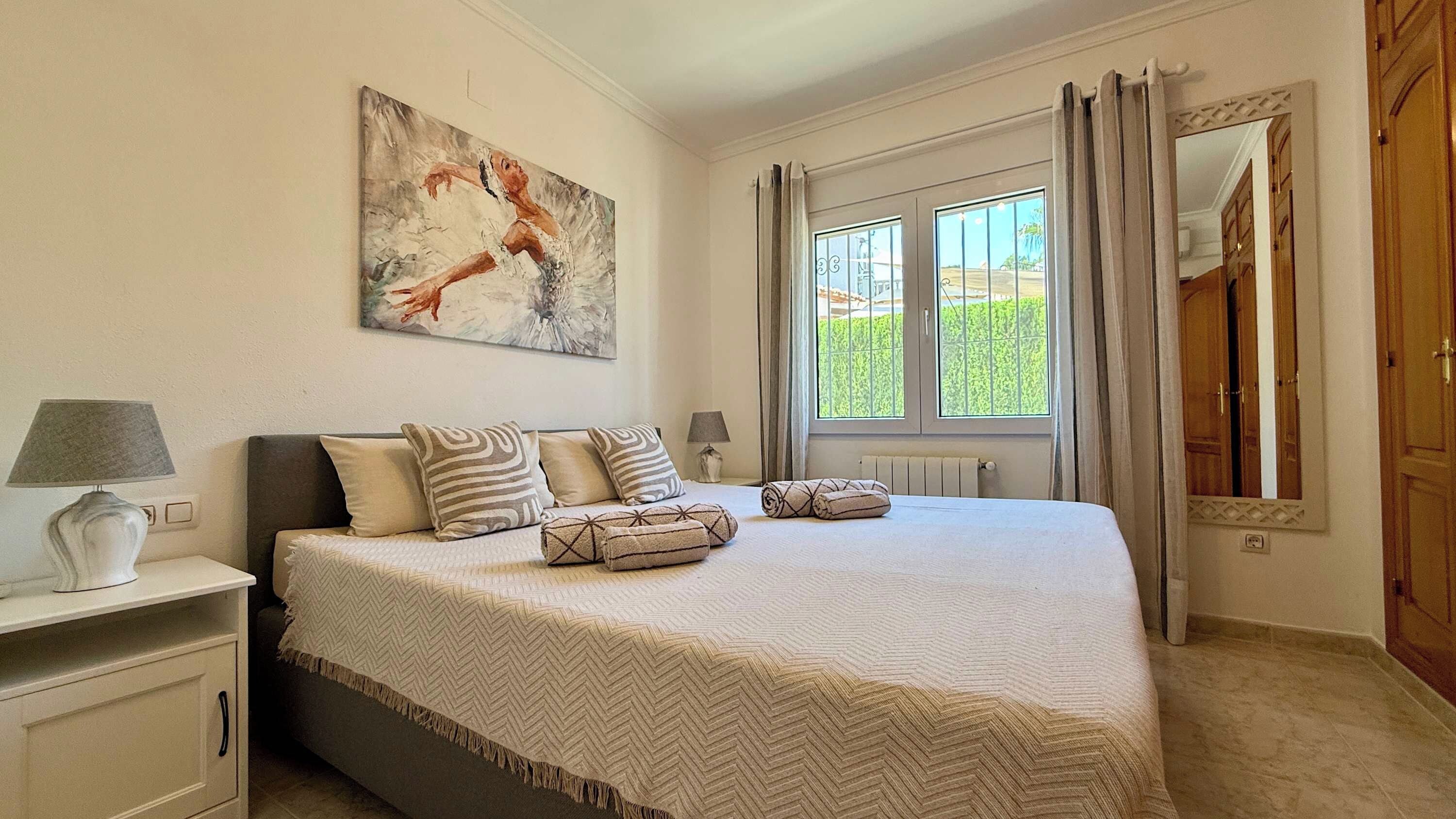 Un dormitorio con cama, mesita de noche, lámpara, cuadro, ventana con cortinas y puerta.