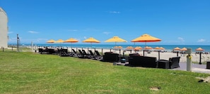 A beachside area with lounge chairs and orange umbrellas, a grassy lawn, and a view of the ocean.