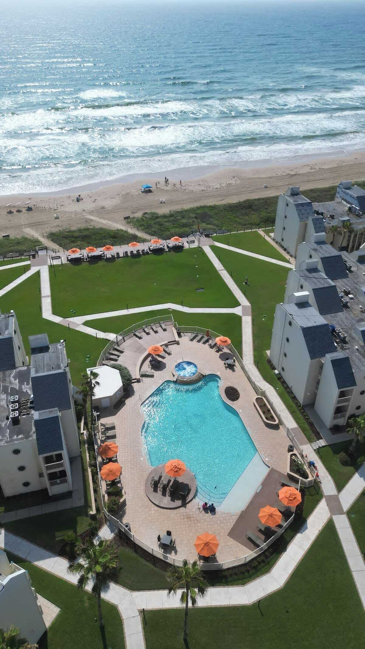 A resort with a central pool surrounded by lounge areas, a beach view, and a well-maintained lawn.