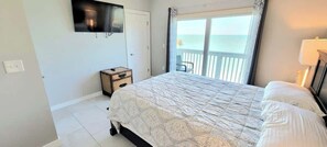A bedroom with a bed, a TV mounted on the wall, a small bedside table, and a window with a view of the sea.